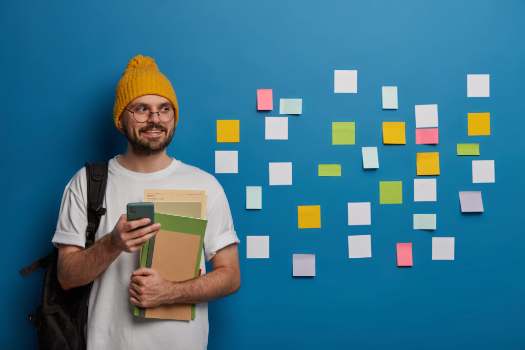 handsome bearded youngster stands with textbooks, mobile phone and rucksack, sends text message to groupmate, glad to pass exam successfully wall with adhesive notes in background. studying, lifestyle handsome bearded youngster stands with textbooks, mobile phone and rucksack, sends text message to groupmate, glad to pass exam successfully wall with adhesive notes in background. studying, lifestyle